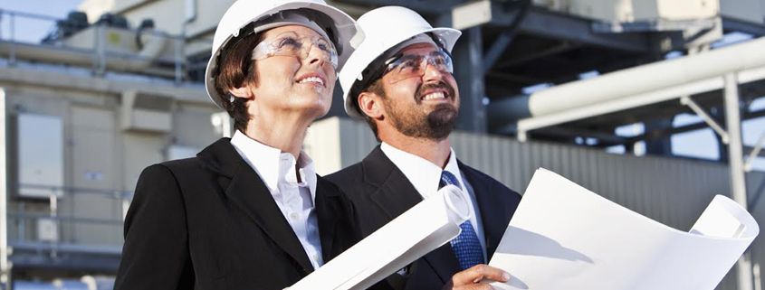 Daytime exterior view of a man and a woman in suits and white hard hats studying blueprints at an industrial site