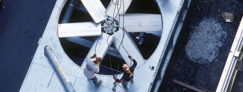 Daytime exterior view of two workers perfroming maintenance on a massive fan in a central heating and cooling system